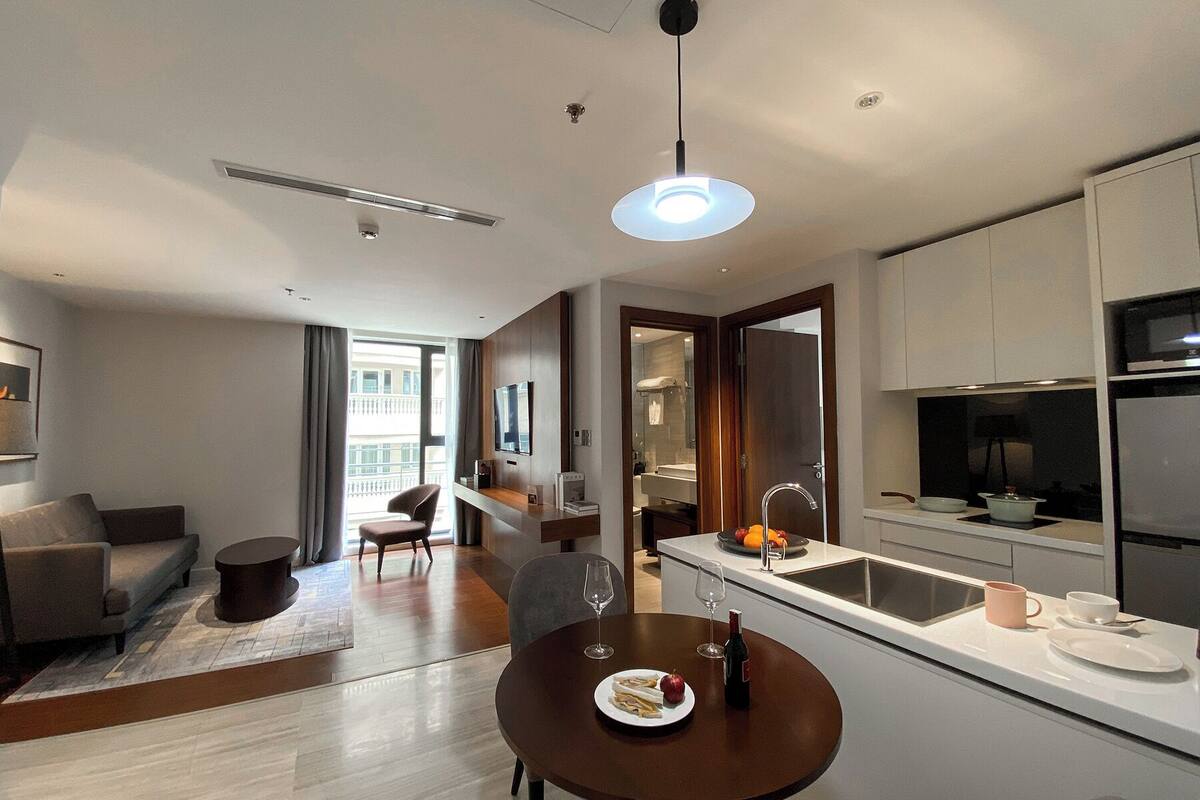 A modern hotel room with a dining area, kitchenette, and a living space with a sofa and chairs.