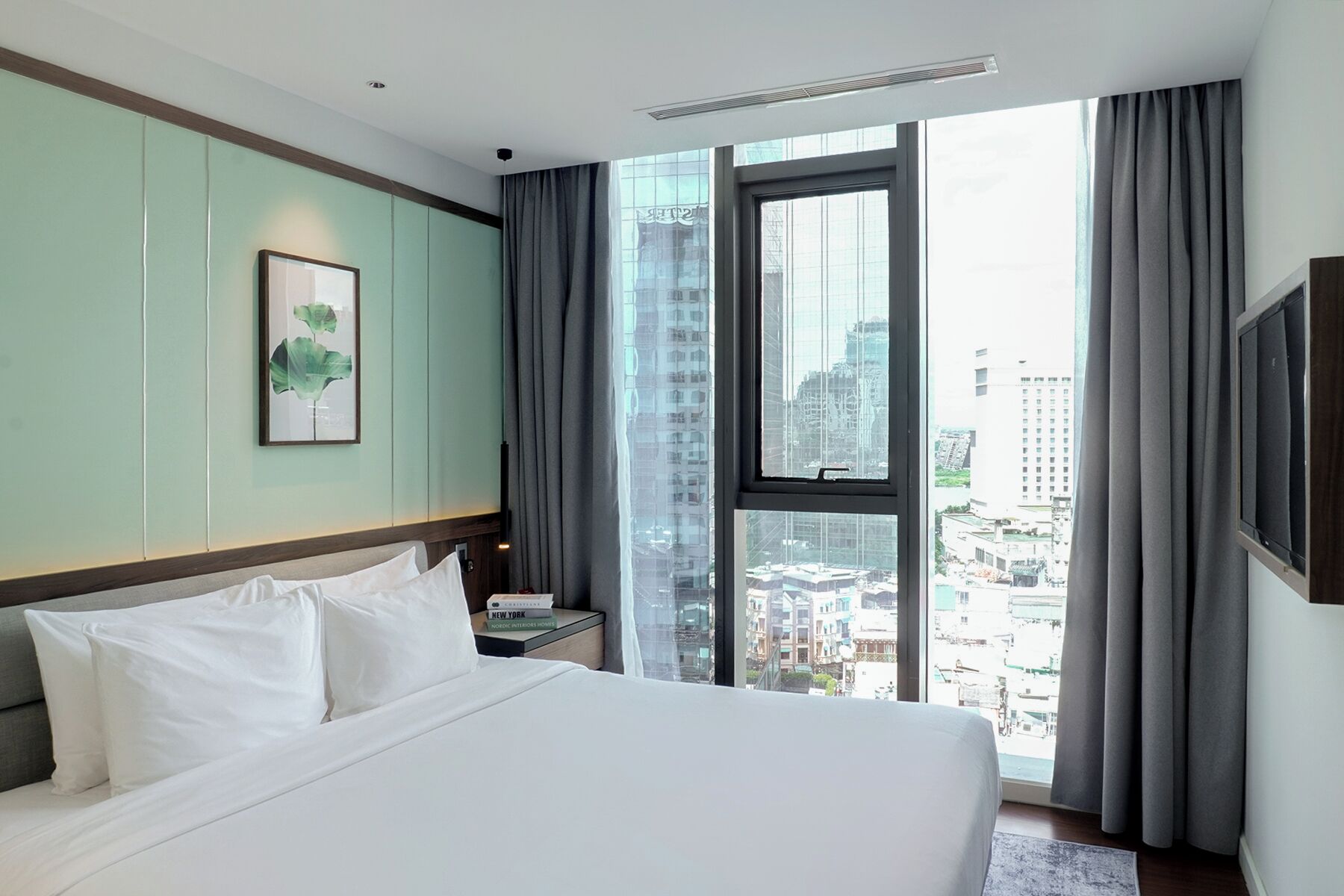 Hotelkamer met een groot bed, een inlijsting foto aan de muur, een nachtkastje met een boek, en uitzicht op de stad door grote ramen.