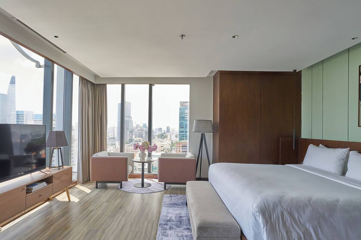 A modern hotel room with a large bed, a TV, a desk, and a view of the city skyline.