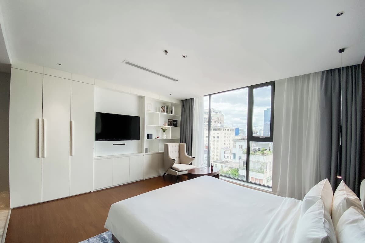 A modern hotel room with a large bed, a flat-screen TV mounted on the wall, a seating area with a chair, and a large window offering a city view.
