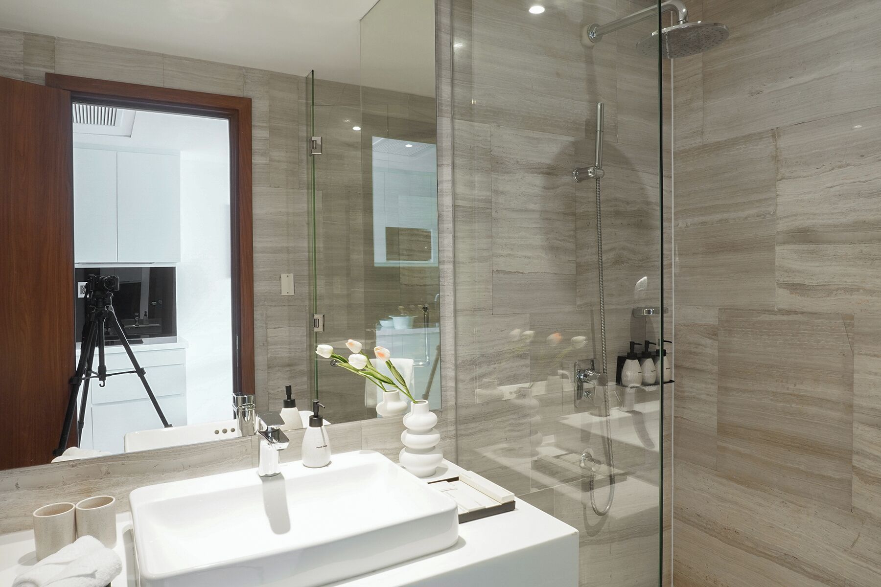 Une salle de bain moderne avec une douche à parois de verre, un lavabo blanc avec deux tasses et un vase rempli de fleurs.