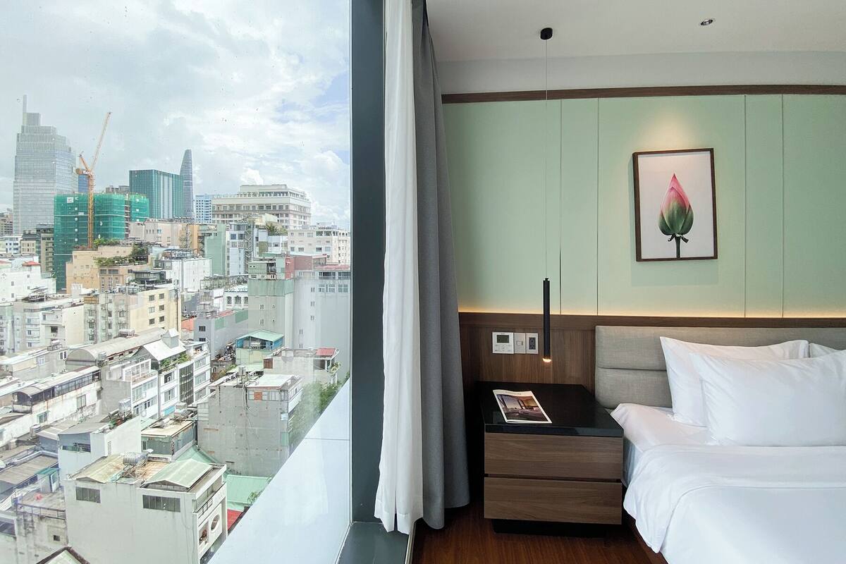 A hotel room with a city view, a bed, a bedside table with a magazine, and a wall-mounted light fixture.