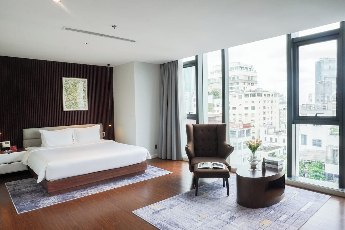 A modern hotel room with a large bed, a comfortable armchair, a small coffee table, and a city view through large windows.