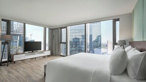 Una habitación de hotel moderna con una cama grande, un televisor sobre una consola de madera, un escritorio y vistas de la ciudad a través de amplias ventanas.