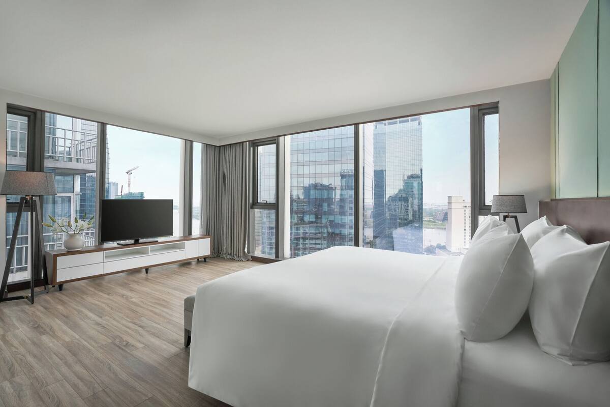 A modern hotel room with a large bed, a TV on a wooden console, a desk, and a view of the cityscape through large windows.