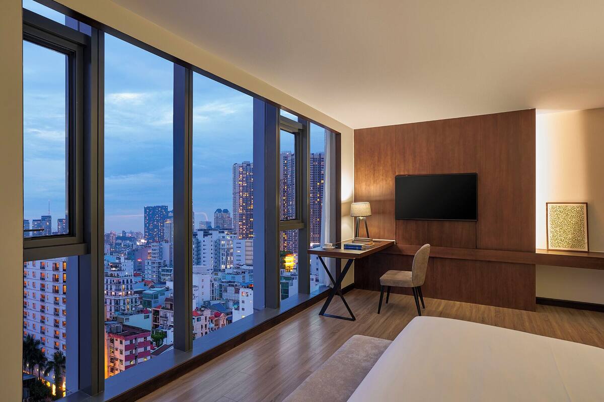 A hotel room with a large window, a desk, a chair, a bed, a flat-screen TV, and a wooden panel wall.