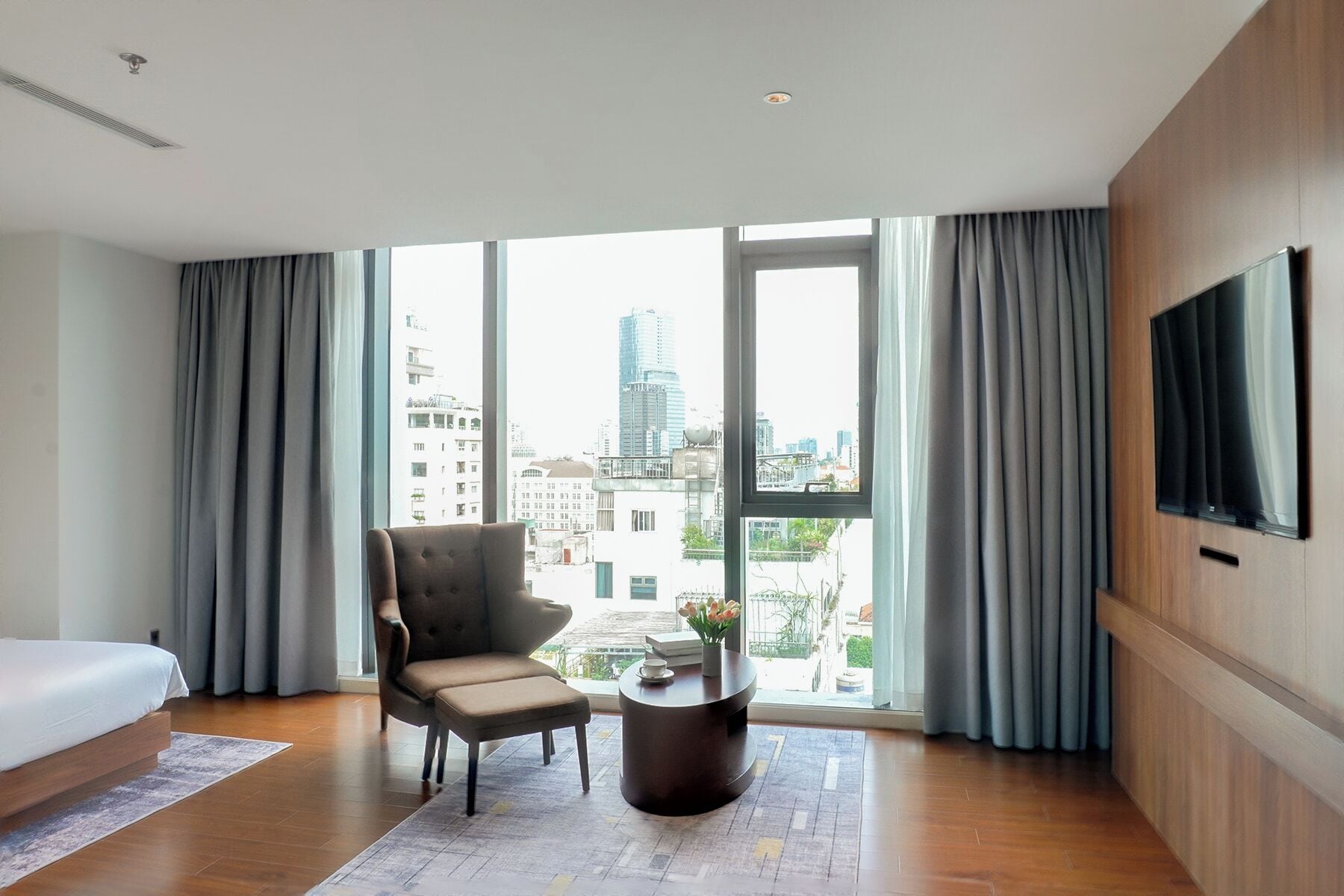 A modern hotel room with a large window, a comfortable armchair, a small round table, and a flat-screen TV mounted on the wall.