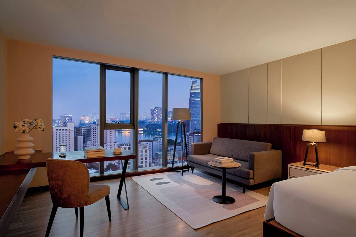 A modern hotel room with a large window offering a city view, a sofa, a small table, and a bed.