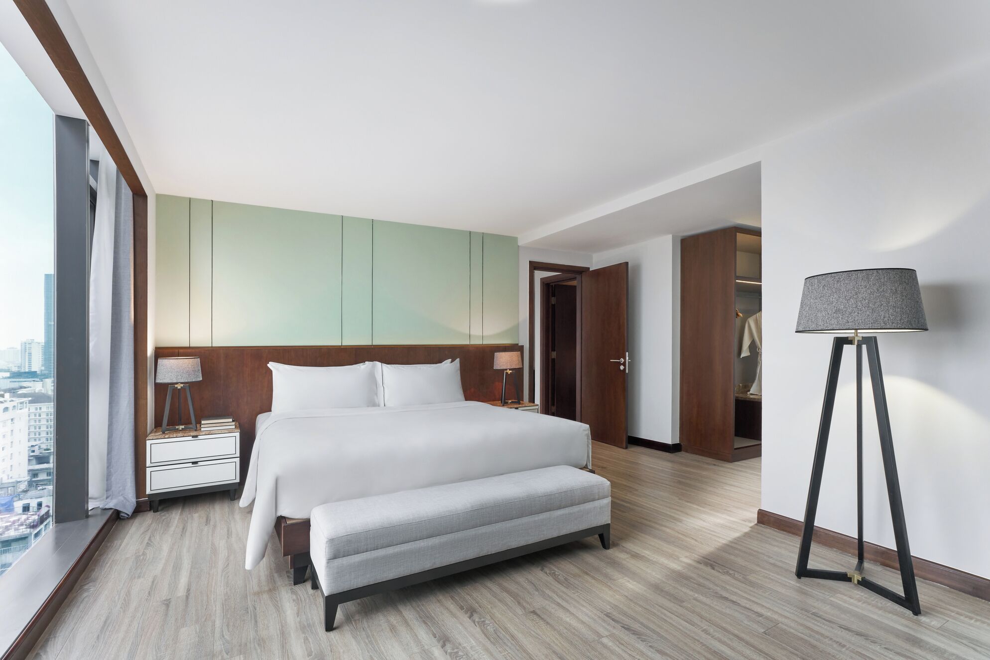 A modern hotel room with a large bed, a bench, a bedside table, a lamp, and a view of the city.