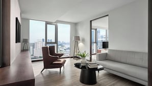 Una sala moderna con sofá, sillón y mesa de centro, ventanas amplias con vista a la ciudad y un televisor de pantalla plana montado en la pared.