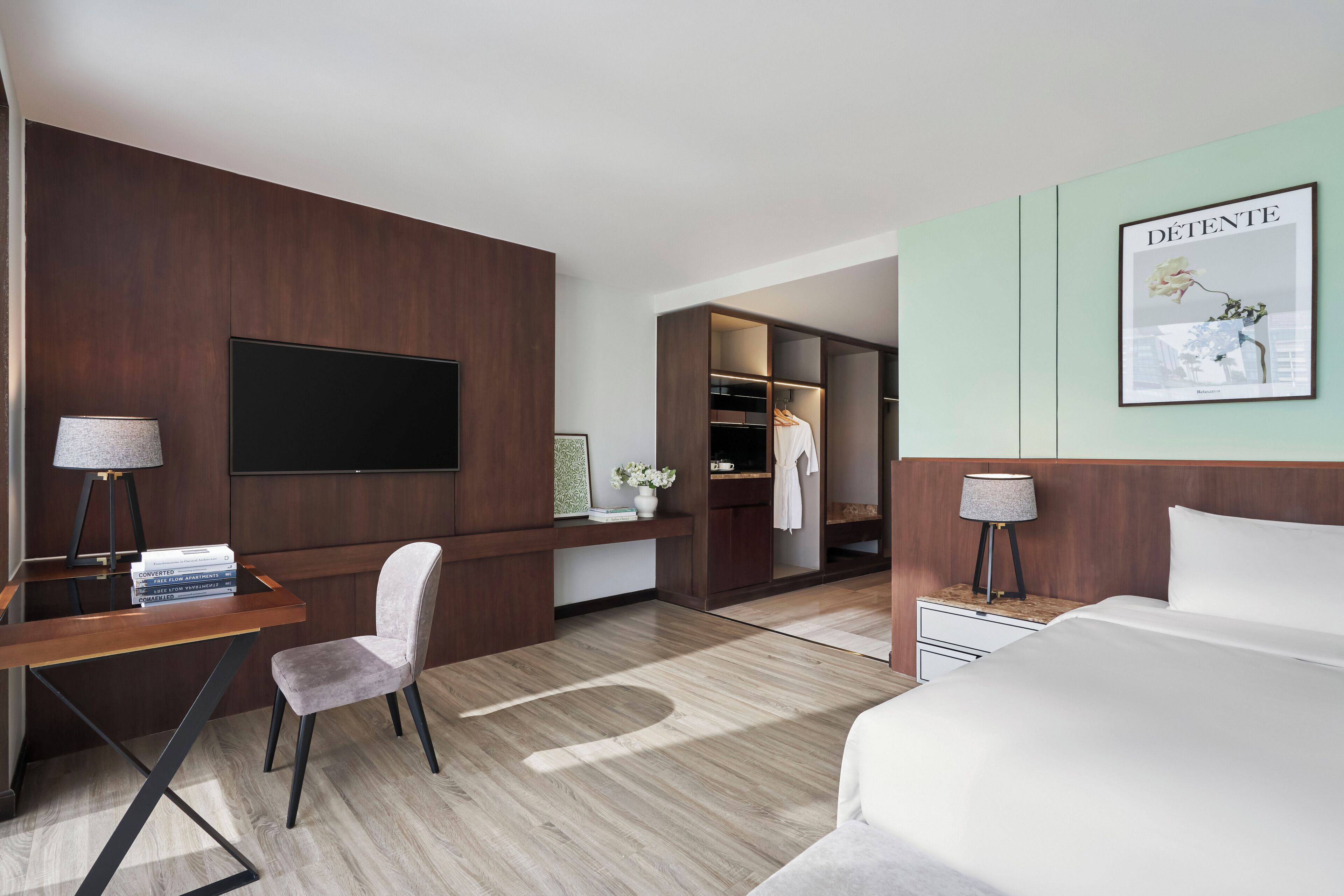 A hotel room with a flat-screen TV, a desk with a chair, a bed, a wardrobe, and a framed picture on the wall.