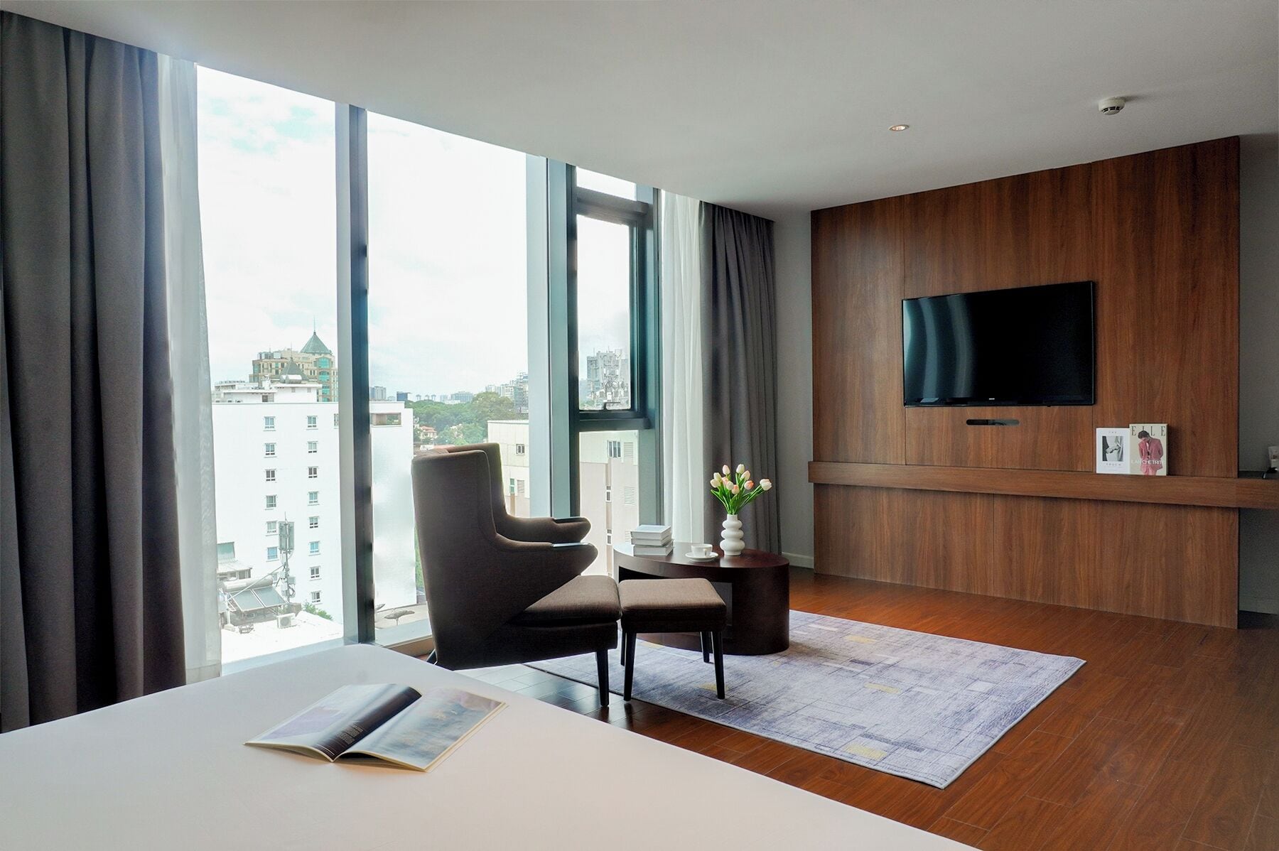 Une chambre d’hôtel moderne dotée d’une grande fenêtre, d’un téléviseur à écran plat fixé au mur, d’un fauteuil confortable, d’une petite table ornée d’un vase de fleurs et d’un lit parfaitement fait.