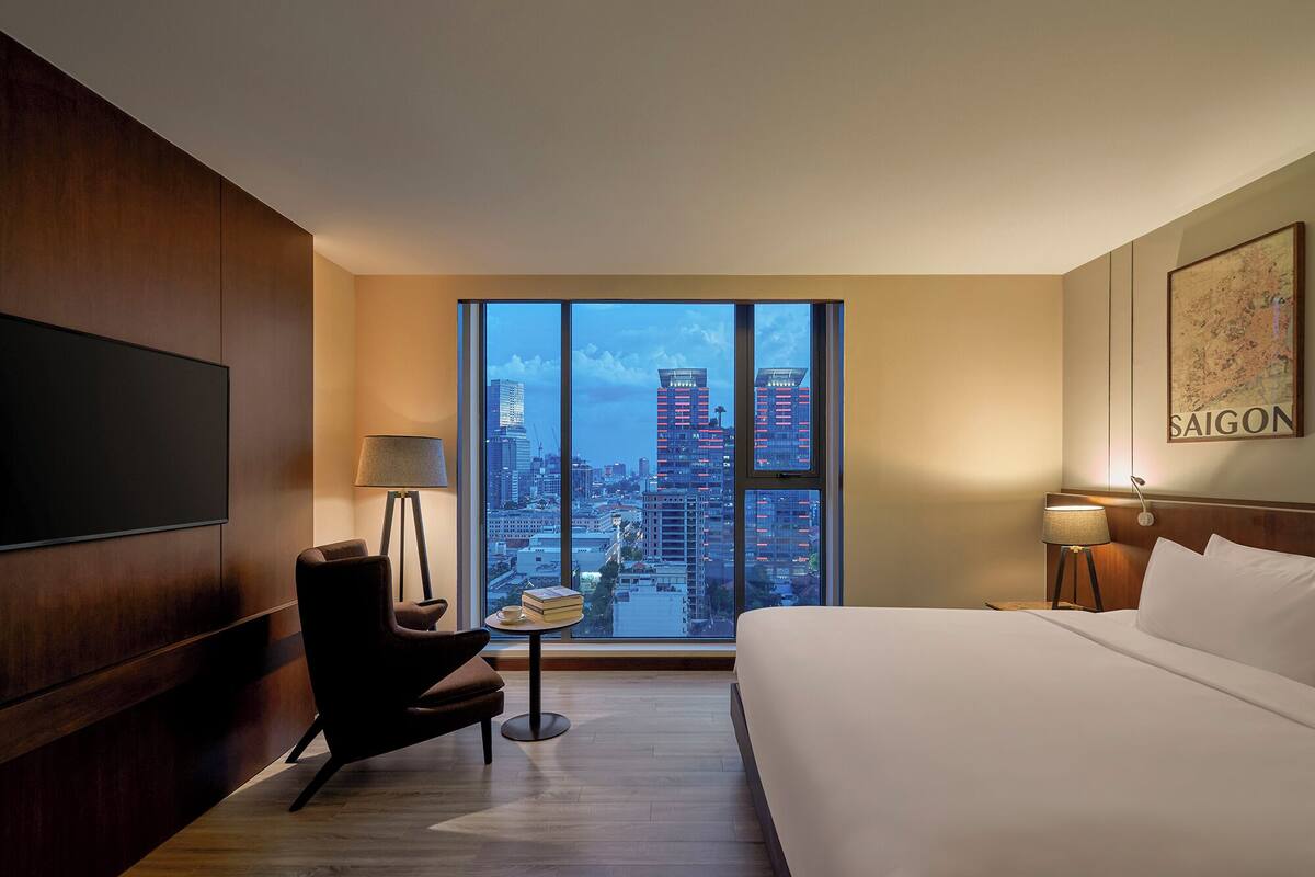 A hotel room with a large window offering a city view, a bed, a chair, a small table with a cup and books, and a flat-screen TV mounted on the wall.