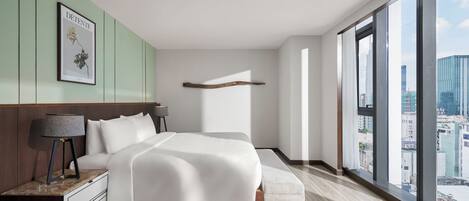 A modern bedroom with a large bed, a nightstand, a bench, and a city view through large windows.