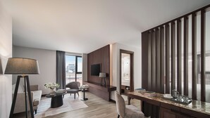 Una habitación de hotel moderna con escritorio, zona de estar y ventanas amplias que dan a vistas de la ciudad.
