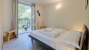 Une chambre d’hôtel moderne avec un grand lit, un banc et une vue sur un jardin, visible à travers une porte vitrée.