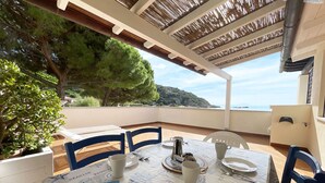 Un espace de restauration extérieur abrité, avec une table dressée pour deux personnes, une vue sur la mer et une pergola au-dessus.