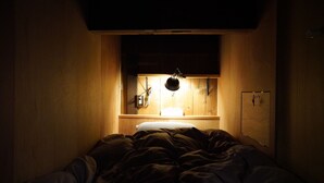 Ett litet, svagt upplyst rum med en säng, ett träskåp och en hylla med en lampa.