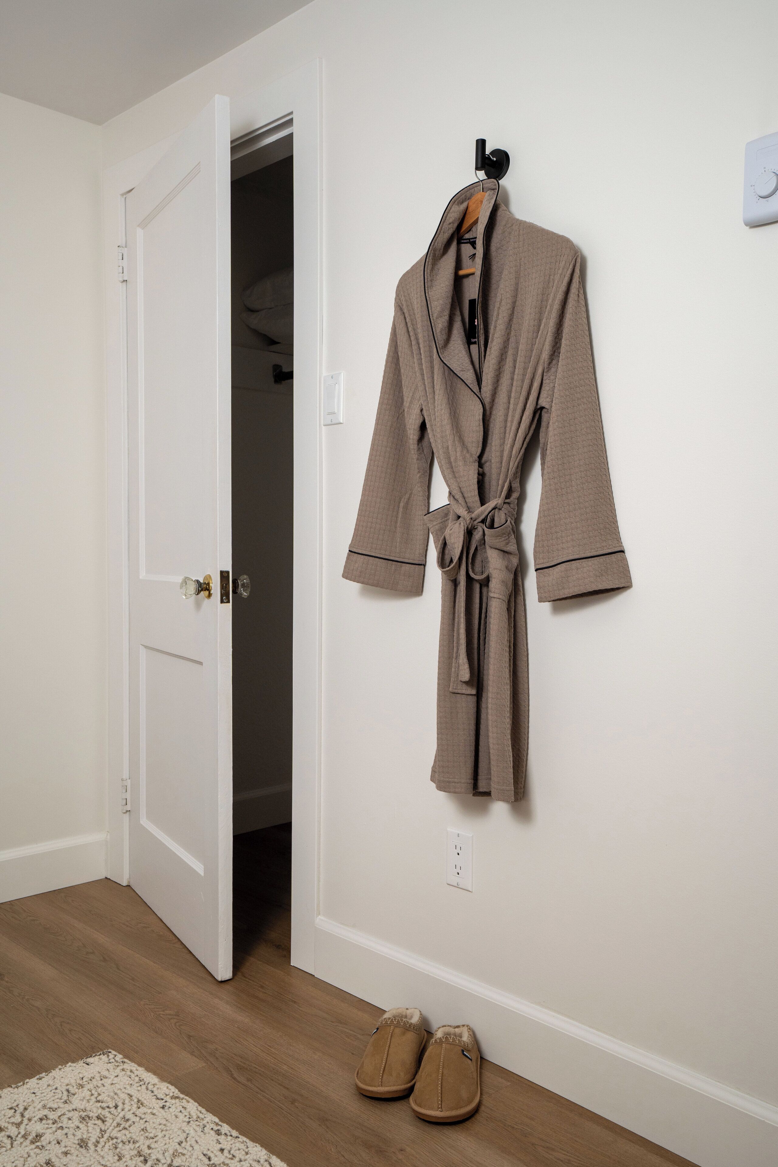 Una habitación con piso de madera, una puerta blanca, una bata beige colgada en la pared y un par de zapatos en el suelo.