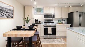 Une cuisine moderne avec des meubles blancs, des appareils en acier inoxydable, une table à manger en bois avec des chaises et une grande fenêtre avec des stores.