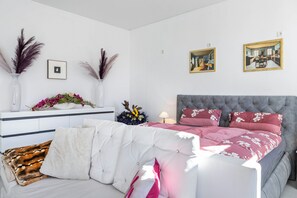 Une chambre à coucher avec une tête de lit capitonnée, un lit recouvert d’une literie à motifs floraux, une table de chevet ornée d’un vase et de fleurs, et deux tableaux encadrés accrochés au mur.