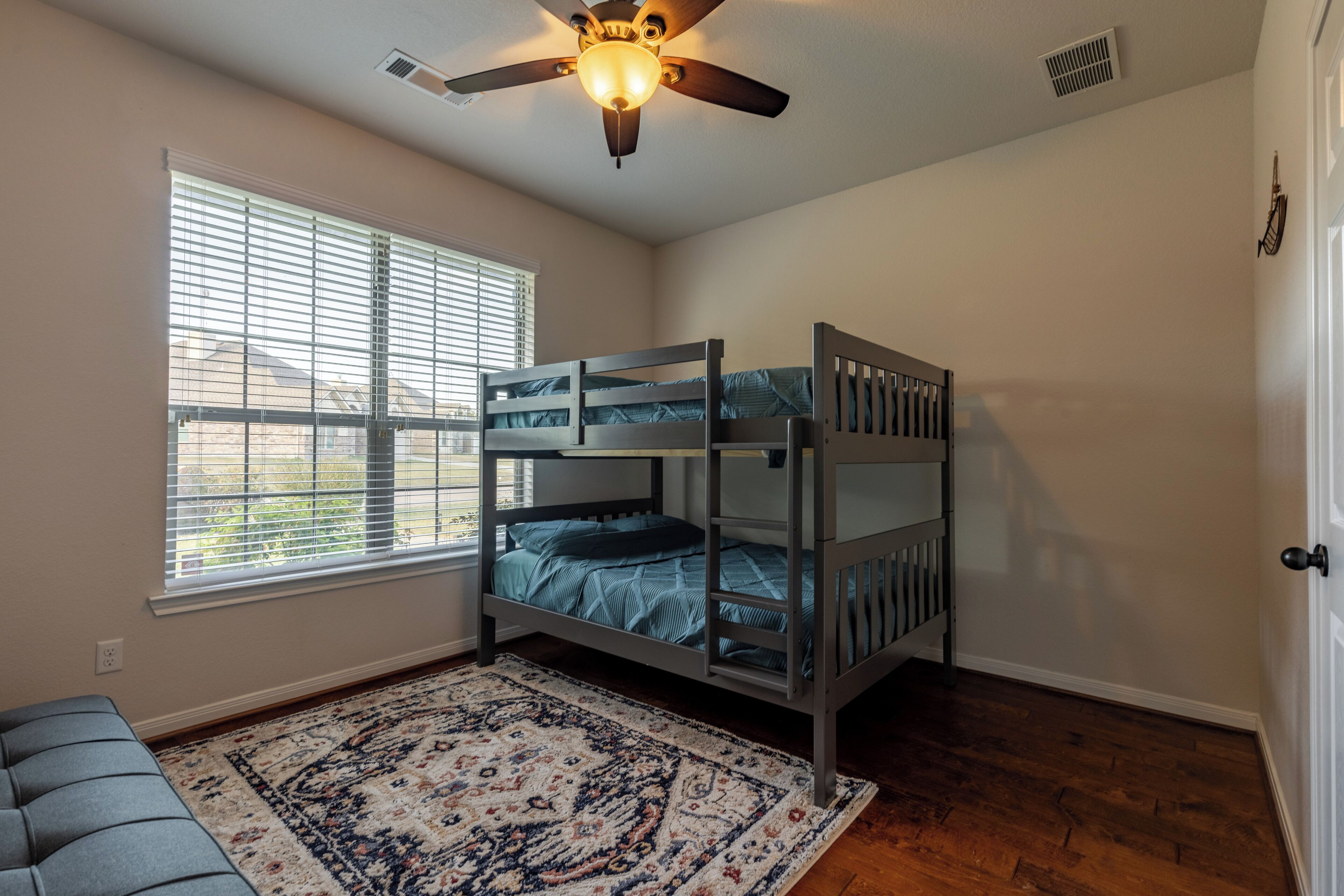 A room with a bunk bed, a ceiling fan, a window with blinds, and a patterned rug.