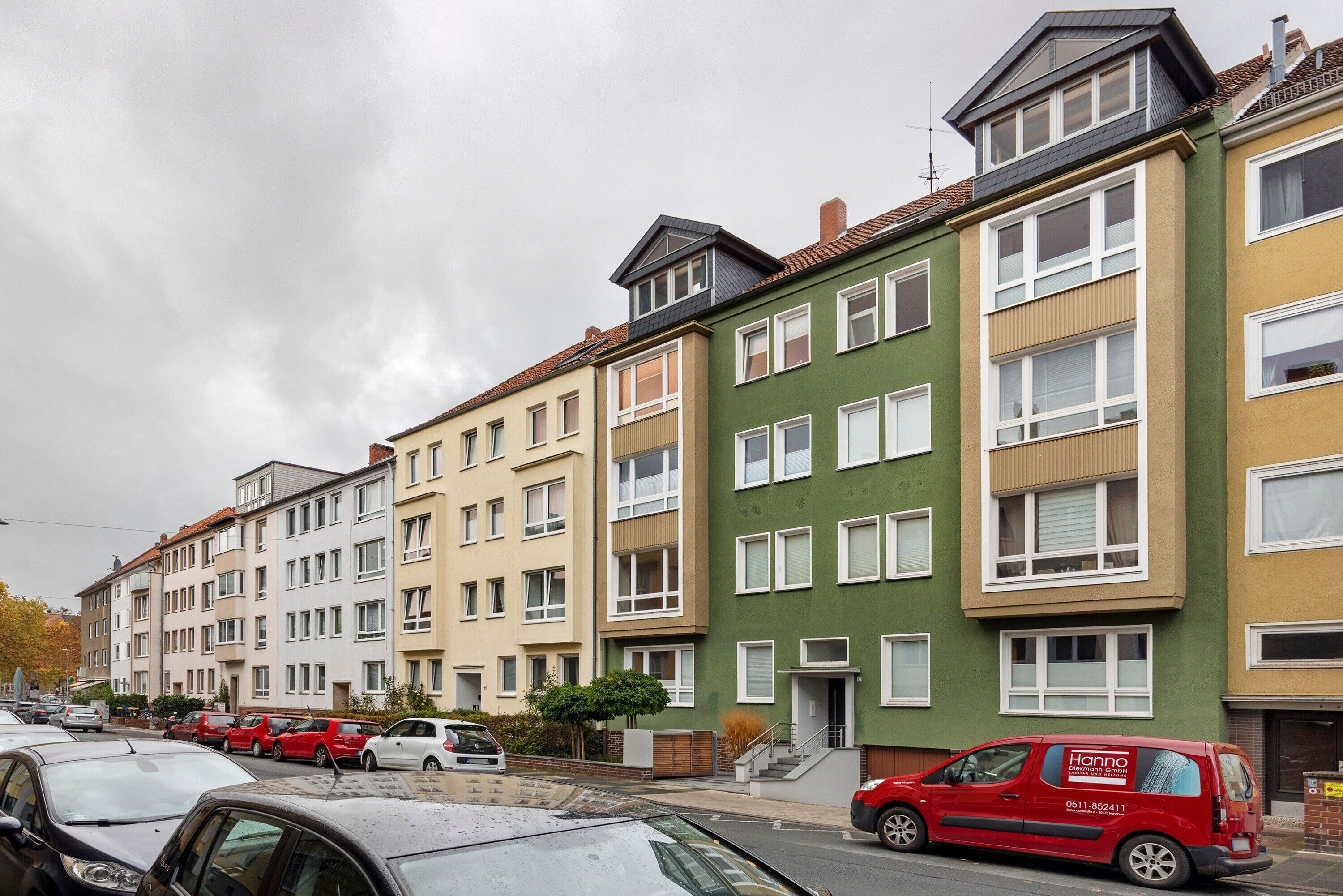 A row of multi-story residential buildings with balconies, a red van parked in front, and cars parked along the street.