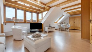 A spacious loft with high ceilings, wooden beams, a flat-screen TV, white furniture, and large windows offering views of the city. - Apartment 'Hannover Südstadt-Perle' with Private Garden and Wi-Fi (Hannover)