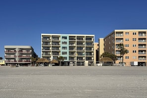 Una hilera de edificios de varios pisos con balcones, suelo arenoso y cielo azul despejado.