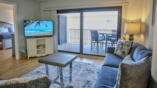 Un salon comprenant un canapé, une table basse, un téléviseur et un balcon aménagé avec du mobilier d’extérieur.
