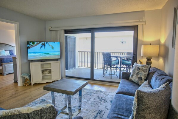A living room with a sofa, a coffee table, a TV, and a balcony with outdoor furniture.