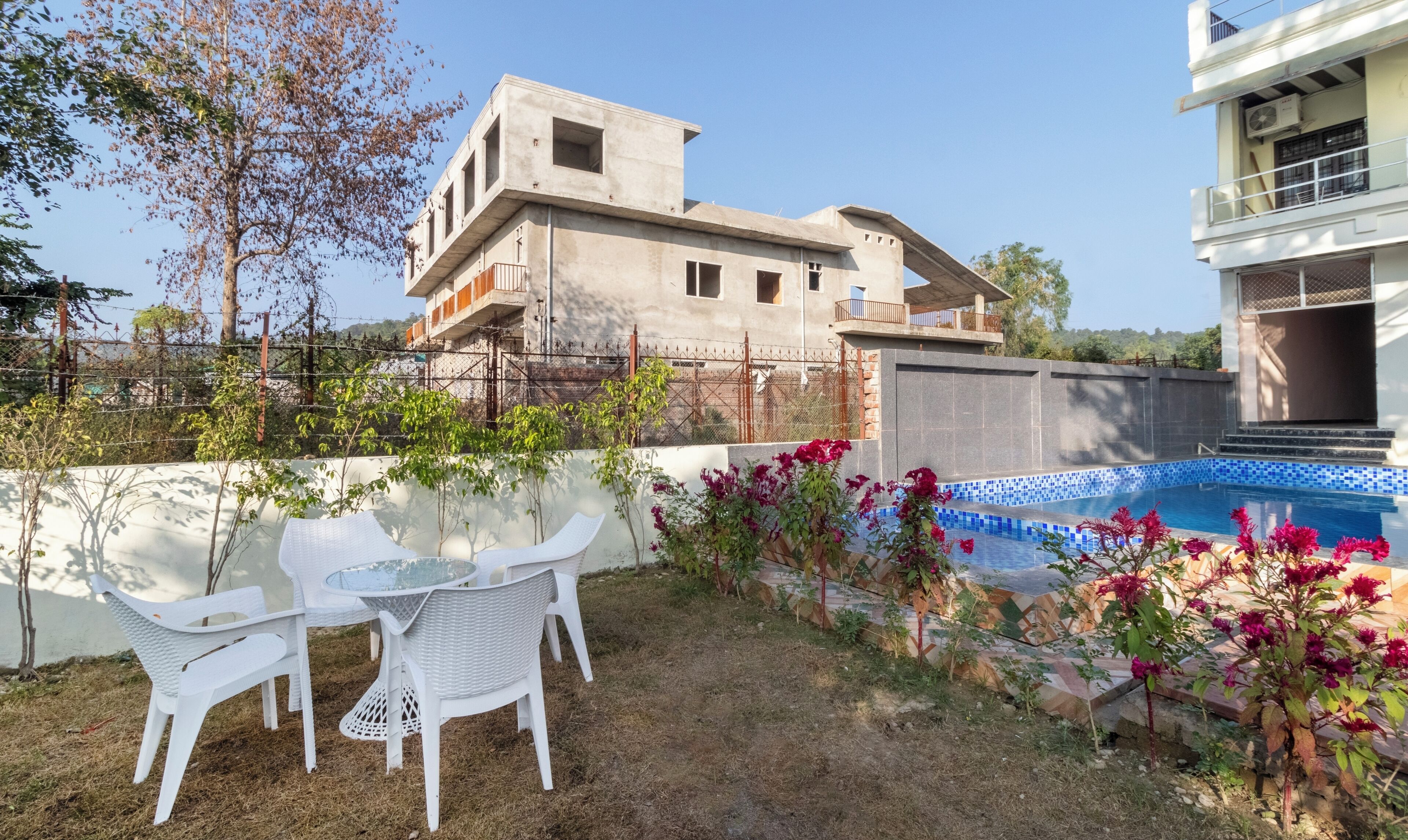 A pool area with a white outdoor table and chairs, surrounded by flowering plants and a view of a building with a balcony.