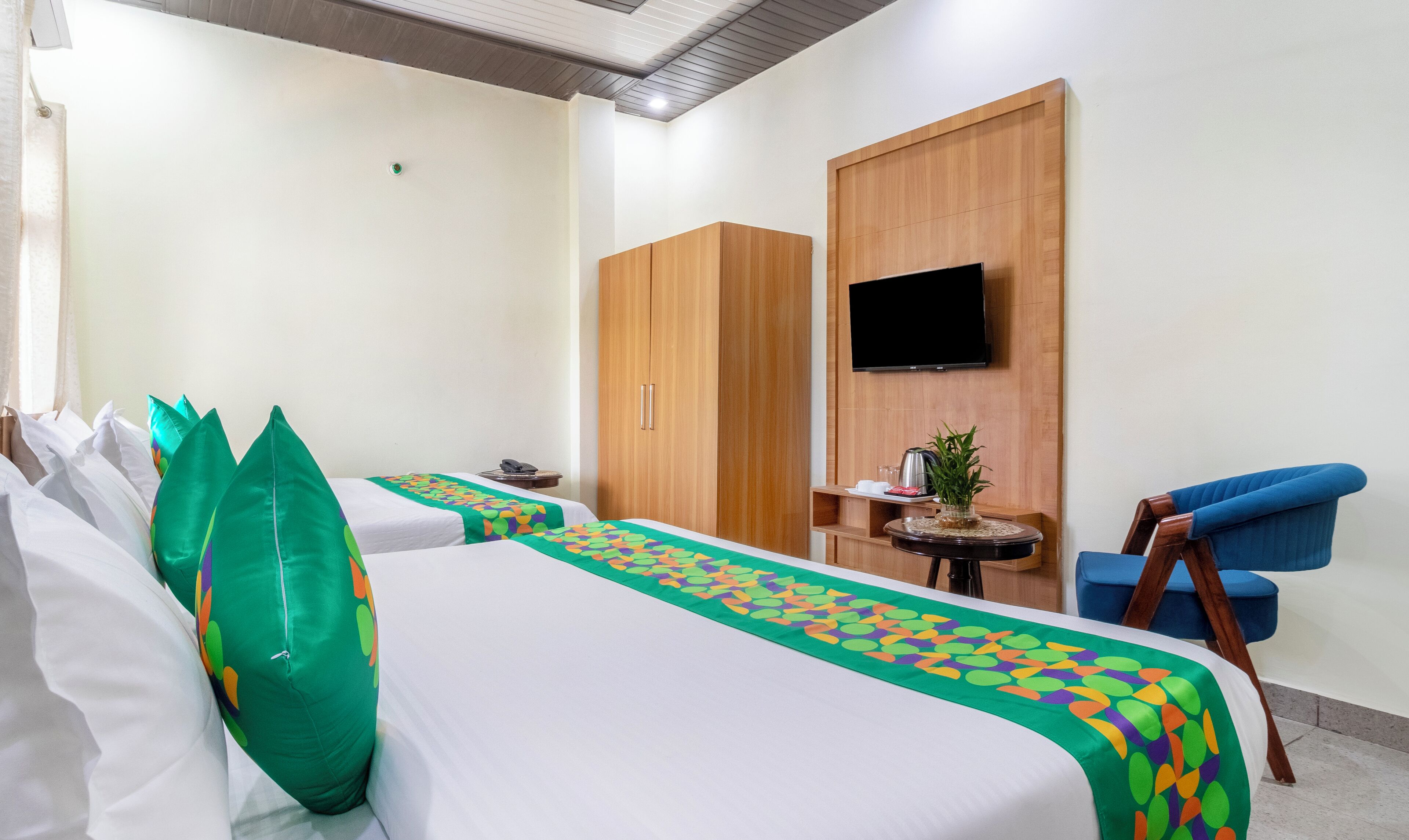 A hotel room with two beds, a TV, a small table with a plant, and a blue chair.
