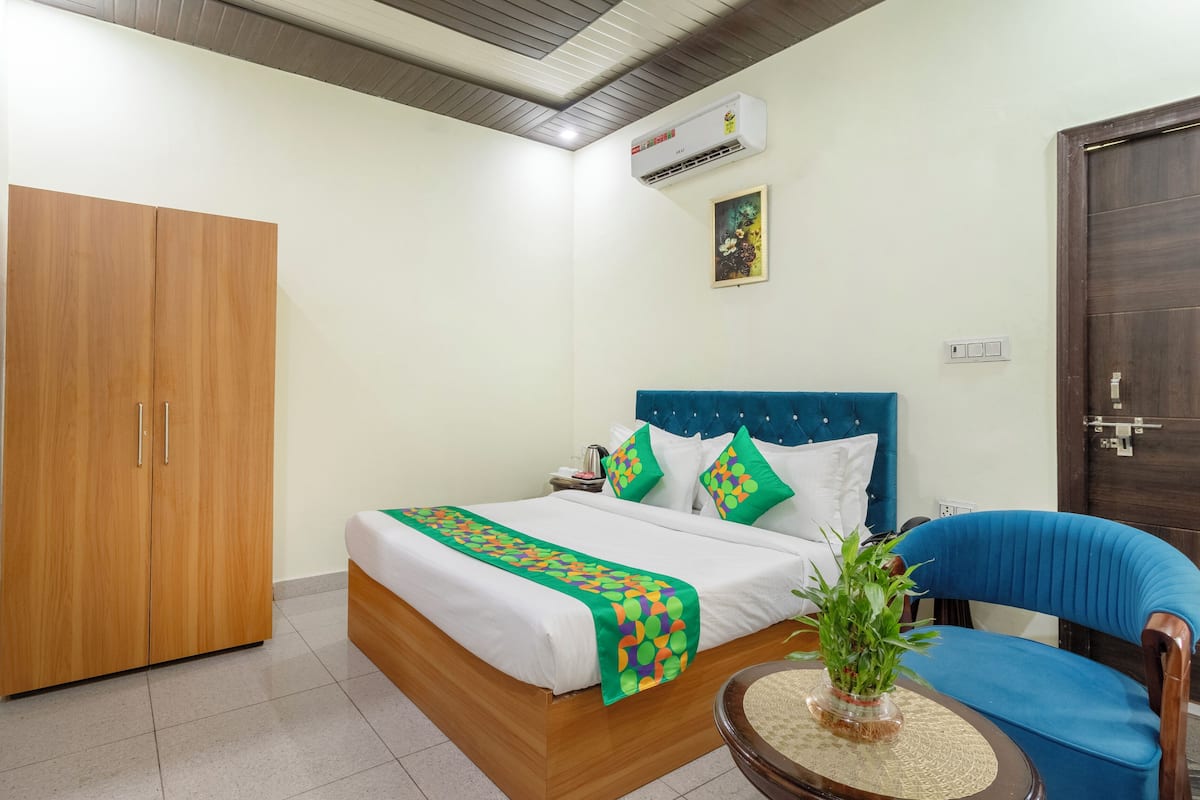 Habitación de hotel con un armario de madera, una cama con cabecera azul, colcha verde y blanca, una silla azul y una mesita con una planta.