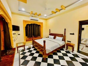 A bedroom with a checkered floor, a wooden bed with white linens, a small bedside table, a wall-mounted TV, and an air conditioning unit. - Hotel Fort Junction Jaisalmer (Jaisalmer)