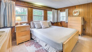 Un dormitorio con pared de paneles de madera, una cama con colcha acolchada, un aparador de madera y una mesita de noche con lámpara.