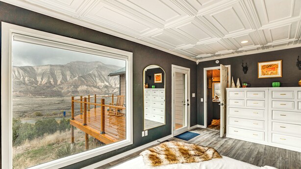 A bedroom with a large window, a wooden deck, a white dresser, and a view of a mountainous landscape. - Colorado River Getaway in Parachute: Dogs Welcome! (Parachute)
