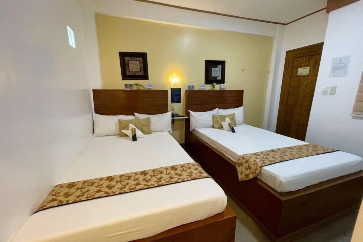 A hotel room with two single beds, each with a wooden headboard, white bedding, and a decorative pillow. There is a nightstand with a lamp between the beds, and a wooden door on the right.