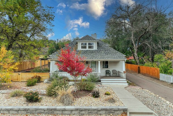 Una casa a un piano con veranda anteriore, un piccolo garage adiacente e un giardino piantumato con diverse piante e un albero dalle foglie rosse.