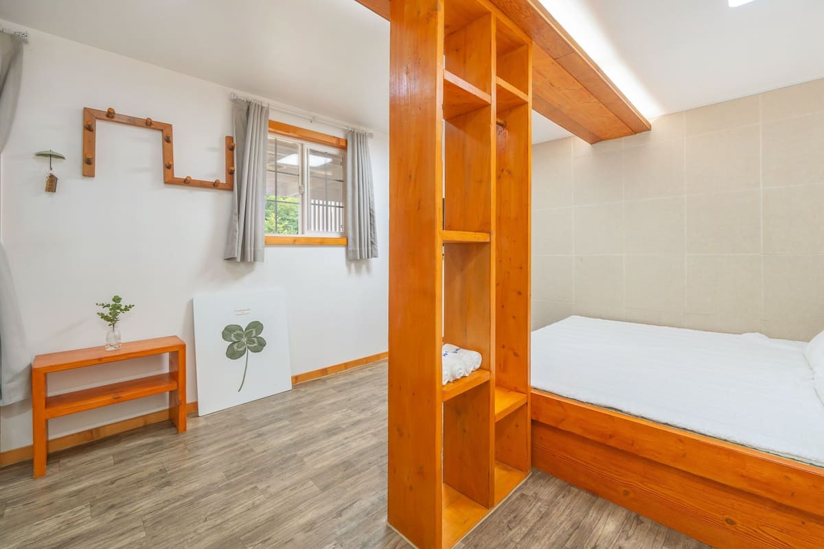 A room with a wooden bed frame, a small bedside table, a window with curtains, and a wall-mounted mirror.