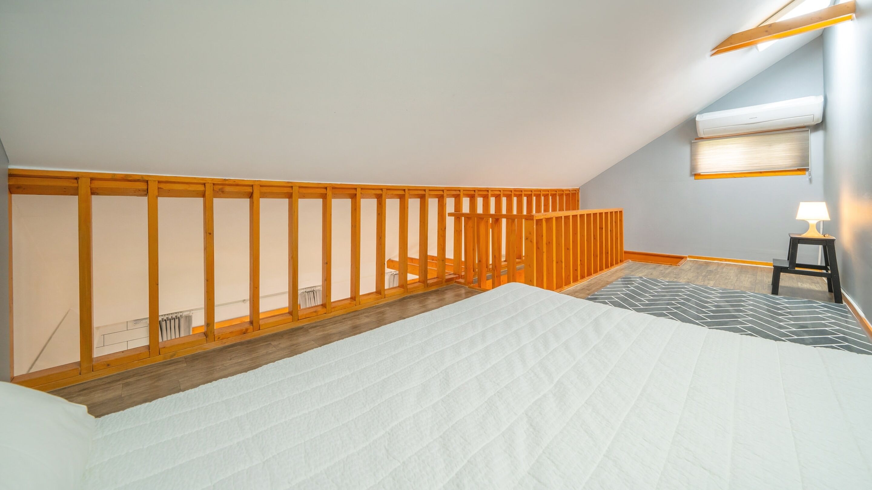 A bedroom with a wooden ceiling structure, a bed with a white quilt, a small table with a lamp, and a radiator on the wall.