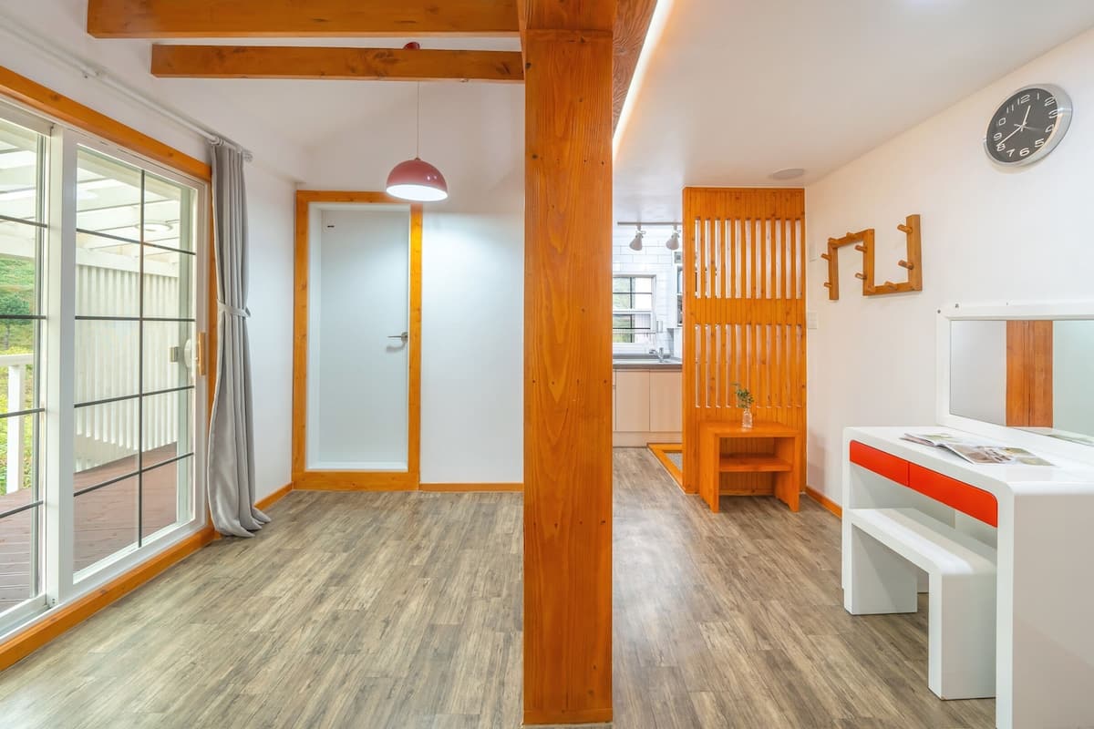 A modern room with a wooden floor, a white desk with a red accent, a large window with a view of greenery, and a wooden partition wall.