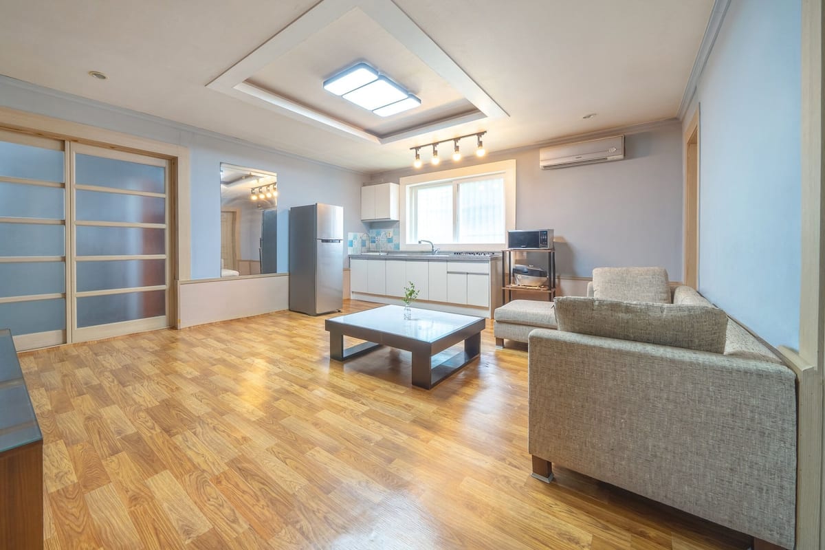 A modern apartment with a wooden floor, a sofa, a coffee table, a TV, a kitchen with a fridge, and a sliding door closet.