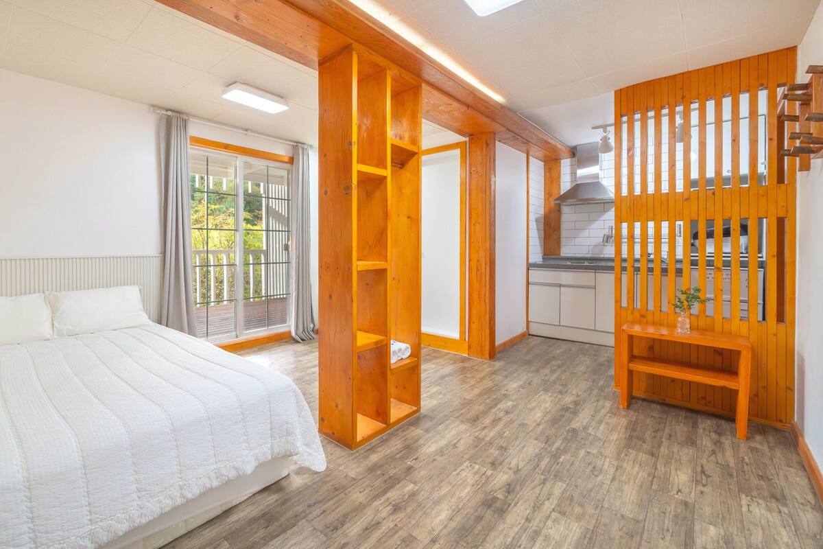 A bedroom with a bed, a wooden wardrobe, a kitchen area, and a window with a view of greenery.