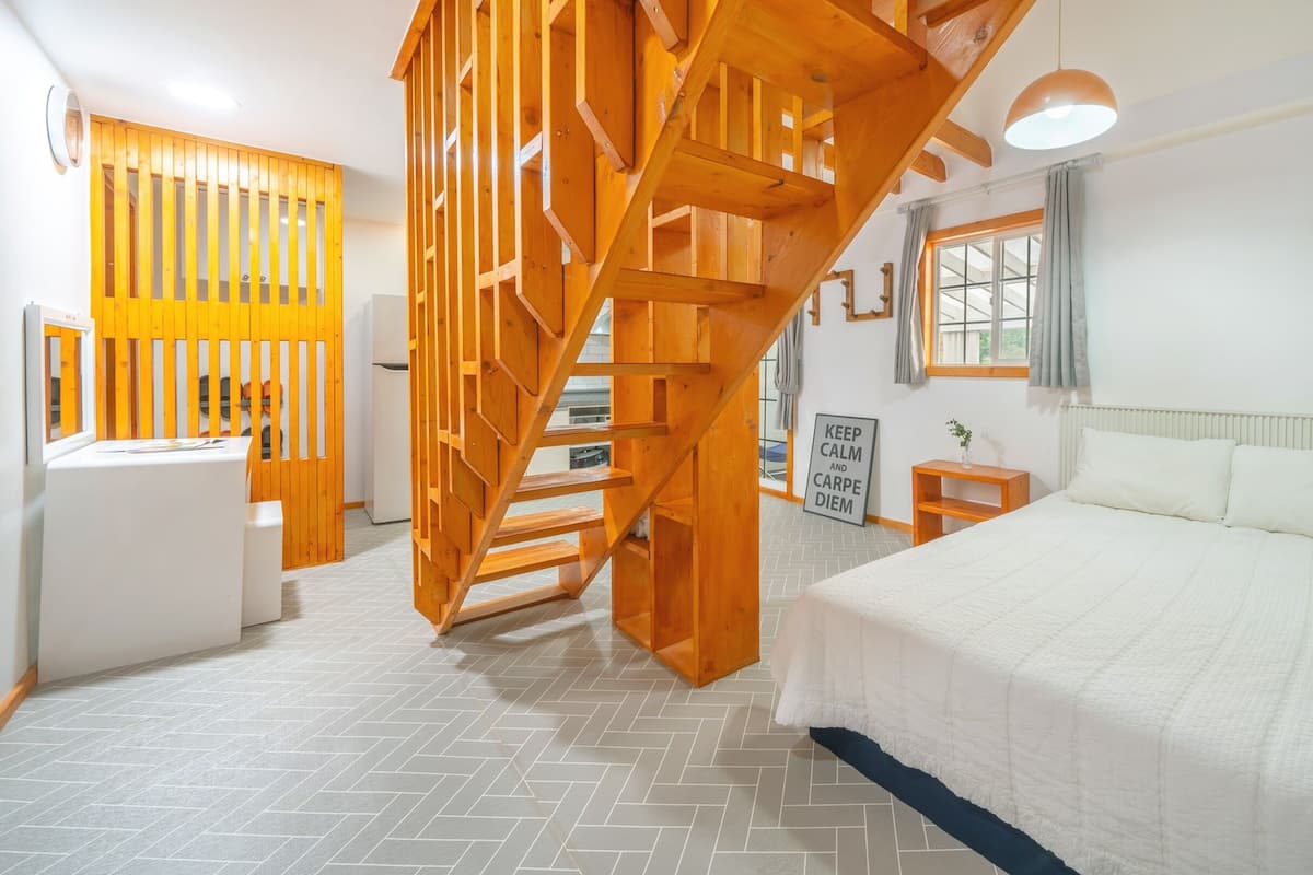 A bedroom with a wooden staircase, a bed, a nightstand, a wall-mounted shelf, and a window with curtains.