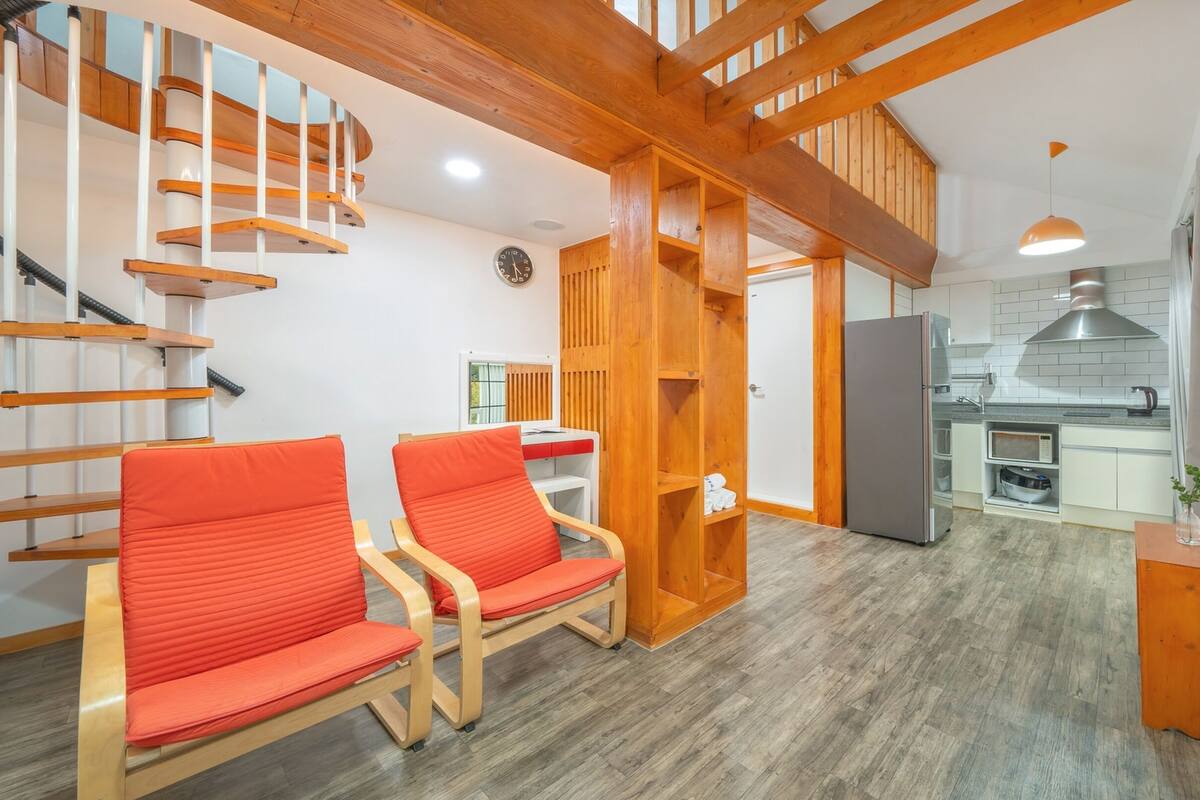 A modern interior with a wooden staircase, two red chairs, a kitchen area, and a small dining table.