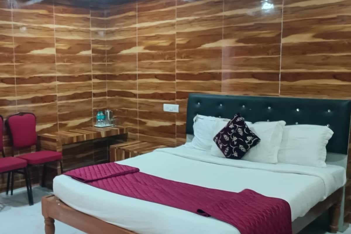 Una habitación con pared de madera, una cama con ropa de cama blanca y una manta roja, dos sillas rojas, una mesita con una botella de agua y una mesita de noche.
