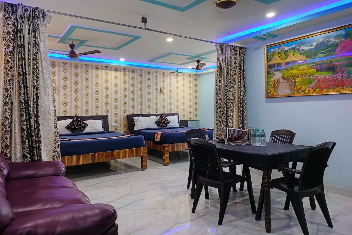Habitación de hotel con dos camas, una mesa de comedor con sillas, un sofá morado, cortinas estampadas y un cuadro de paisaje en la pared.
