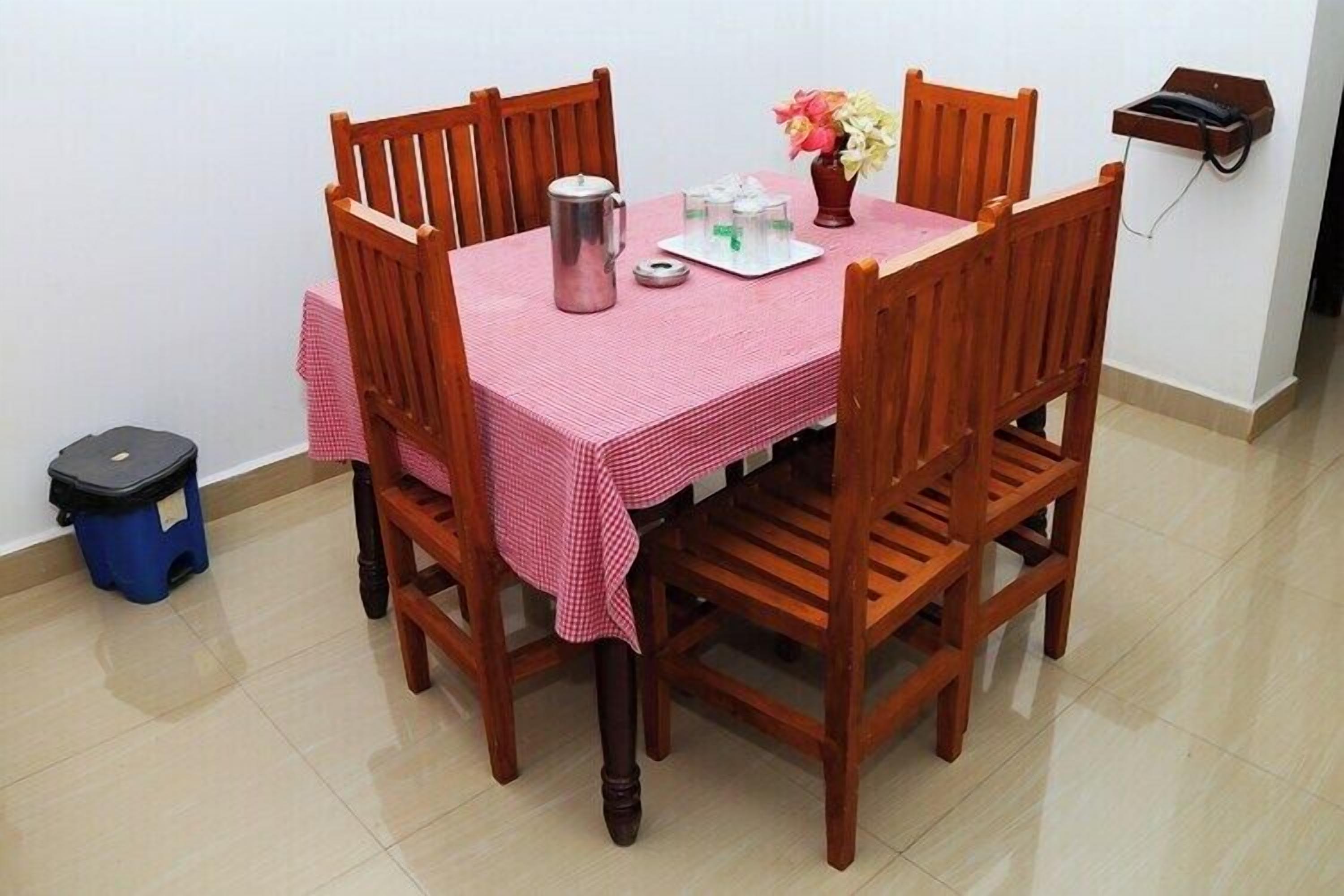 Una mesa de comedor con mantel a cuadros rosa, rodeada de sillas de madera. Hay una jarra metálica y una bandeja con tazas sobre la mesa. También se observa un jarrón con flores.