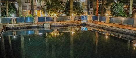A nighttime swimming pool with a tiled deck, surrounded by palm trees and buildings.